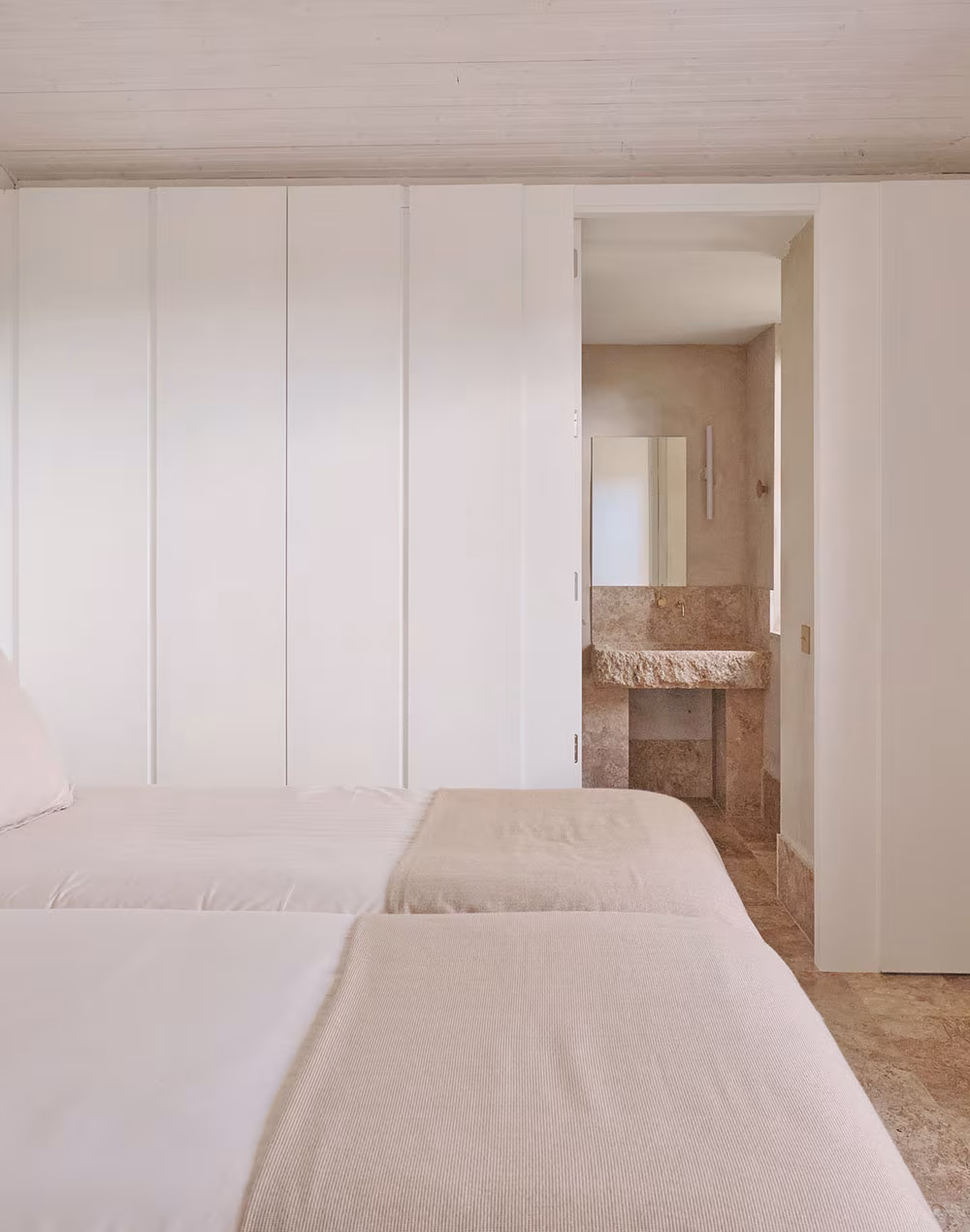 Dormitorio con camas gemelas vestidas en tonos neutros y vista a baño rústico con lavamanos de piedra y grifería dorada.