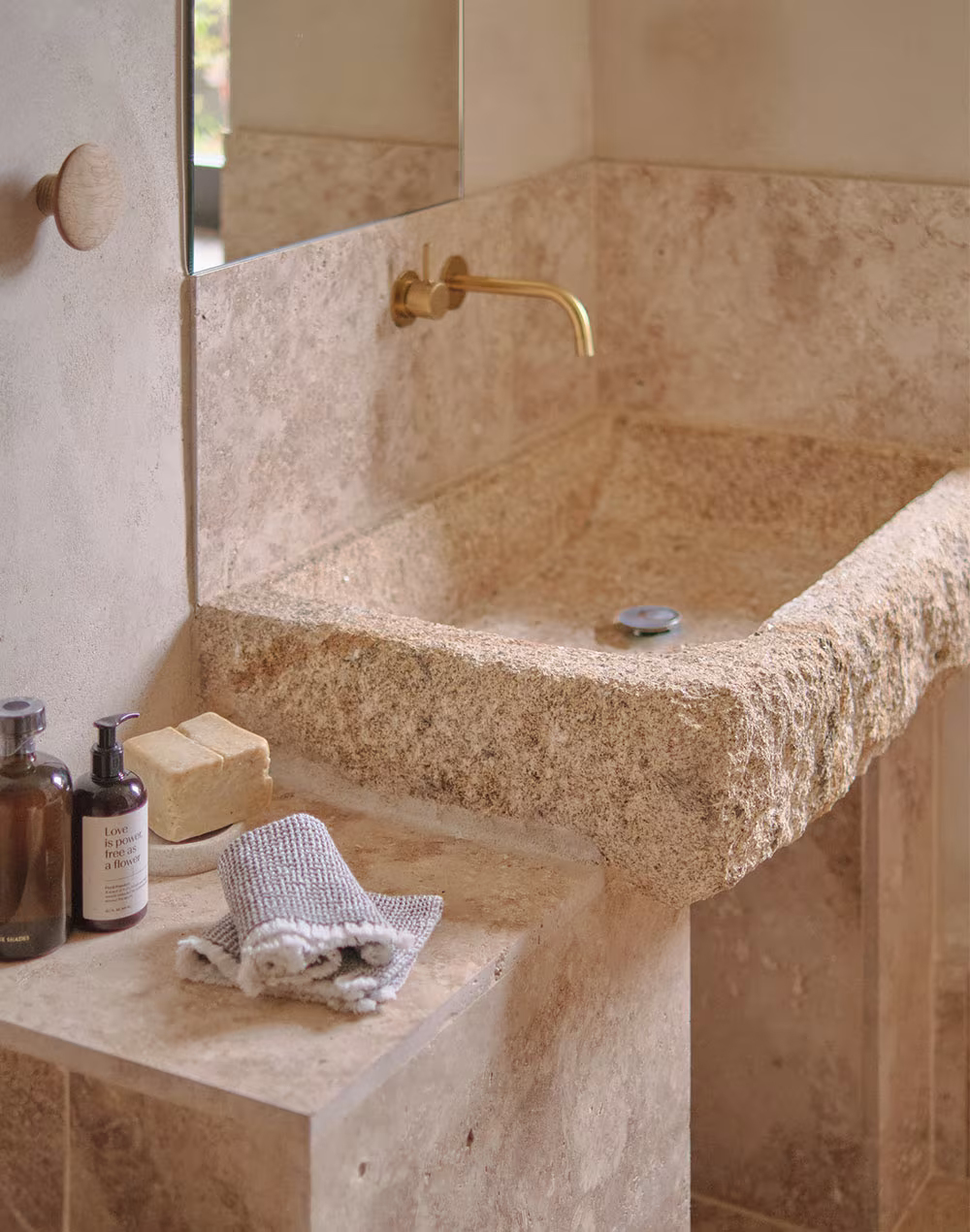 Lavabo rústico de piedra tallada con grifo dorado, espejo rectangular y artículos de tocador sobre encimera de piedra beige.