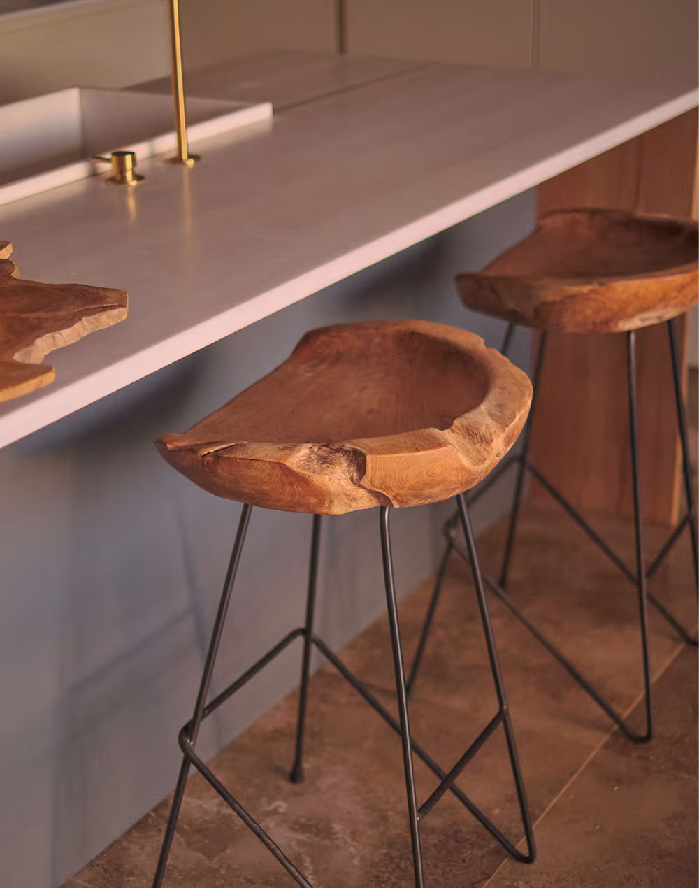 Taburetes altos con asiento de madera natural tallada y patas metálicas negras junto a una barra de cocina blanca con grifo dorado