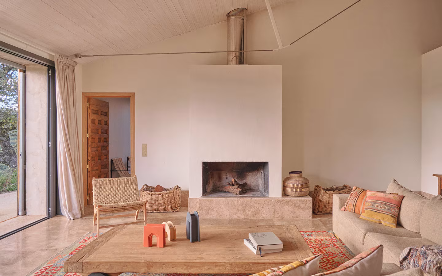 Chimenea blanca al centro con leña, sillas tejidas, sofá con cojines étnicos y luz natural entrando por ventanal a la izquierda.