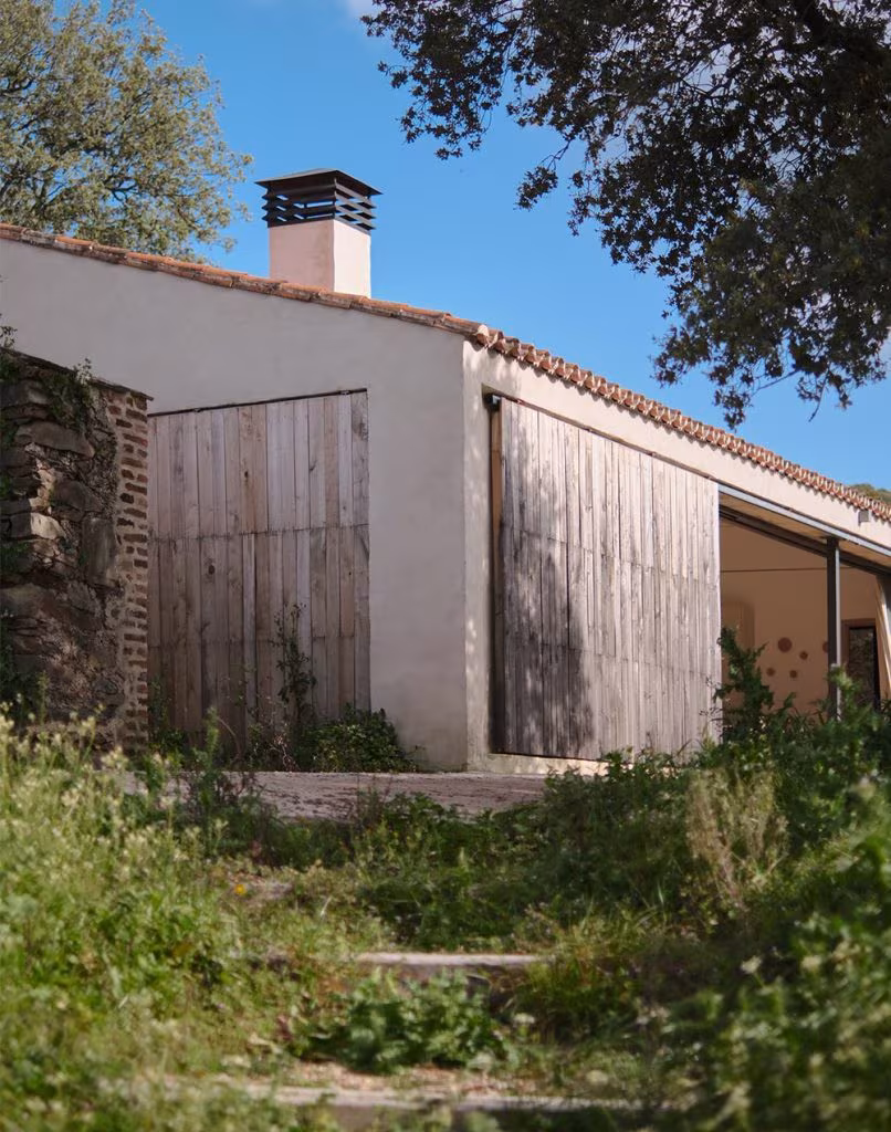 Fachada lateral de casa rural con paredes blancas, tejado a dos aguas y portones de madera, rodeada de vegetación silvestre