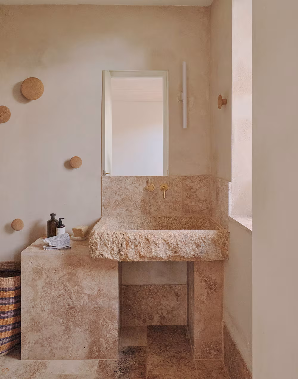 Baño de estilo rústico con lavamanos de piedra tallada, grifería dorada y pared en tono beige con percheros de madera redondos.