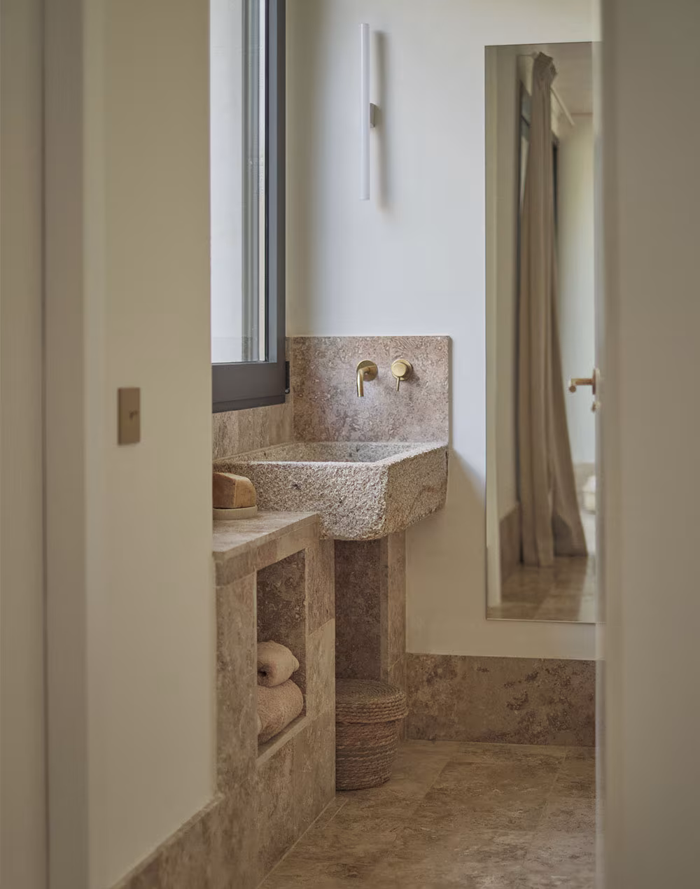 Baño moderno con lavabo de piedra y grifería dorada, iluminación natural, espejo grande en el lateral y decoración minimalista en tonos piedra