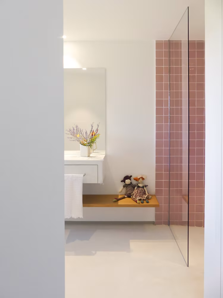 Interior de baño con lavabo blanco, espejo, ducha con mampara de cristal y toques de color rosa y madera.