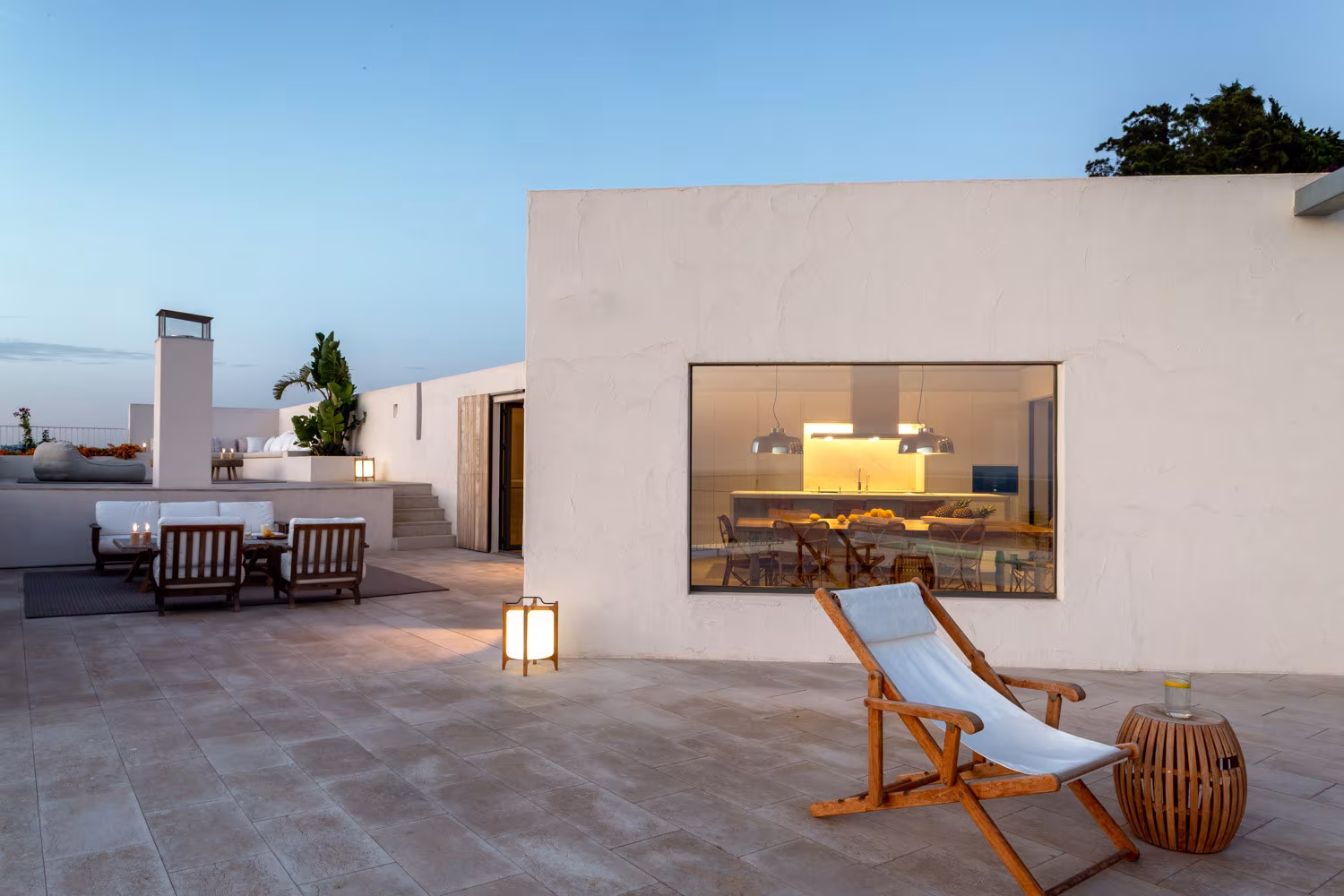 Terraza mediterránea al atardecer con suelo de piedra, sillones de exterior y tumbona de madera