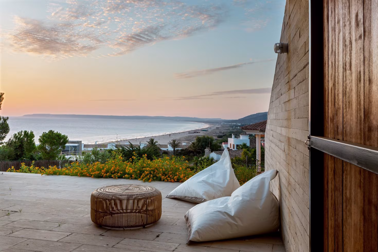 Terraza al atardecer con dos pufs blancos y mesa baja de mimbre sobre suelo de piedra, frente al mar