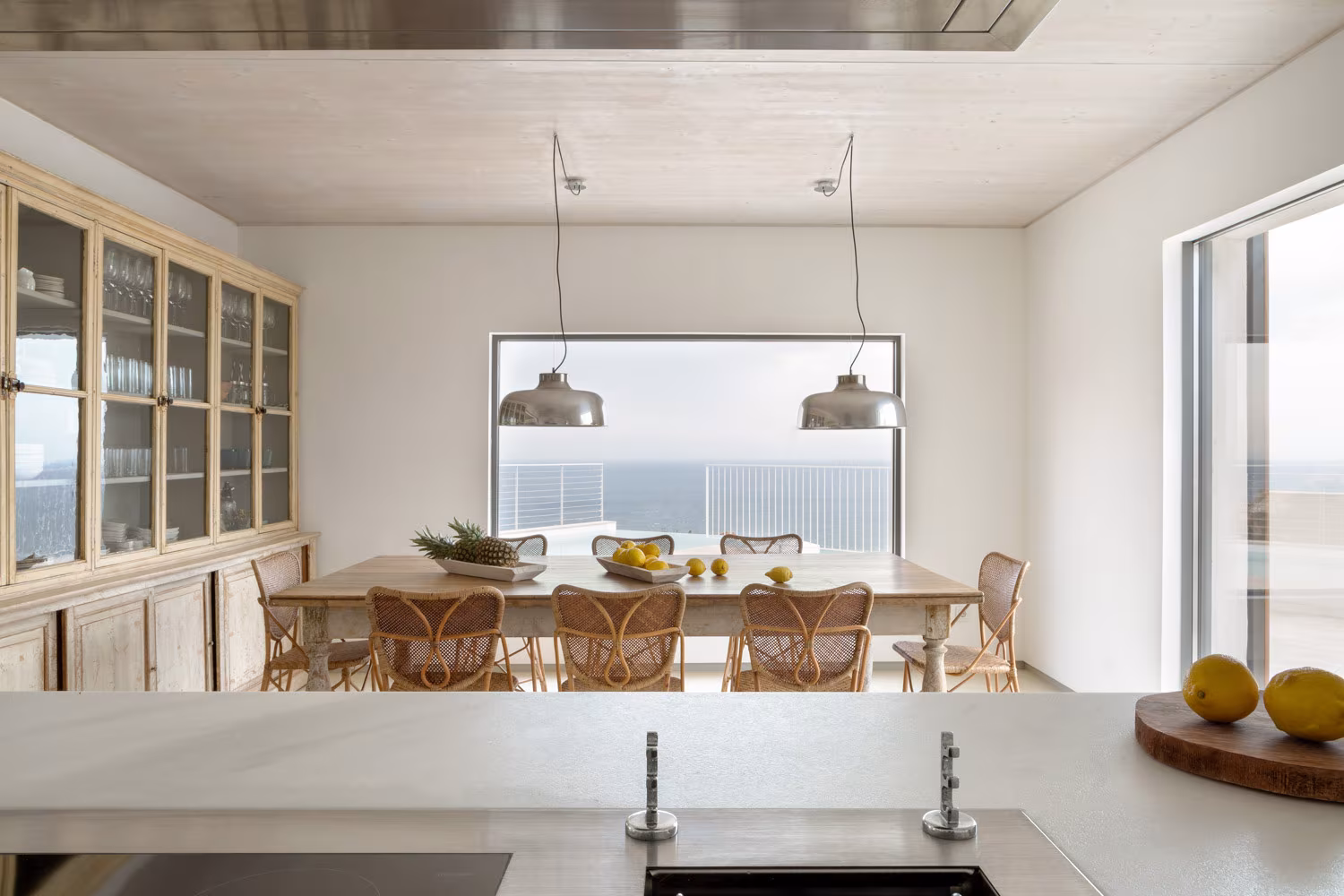 Cocina-comedor mediterránea con isla blanca, mesa rústica y sillas de mimbre frente al mar