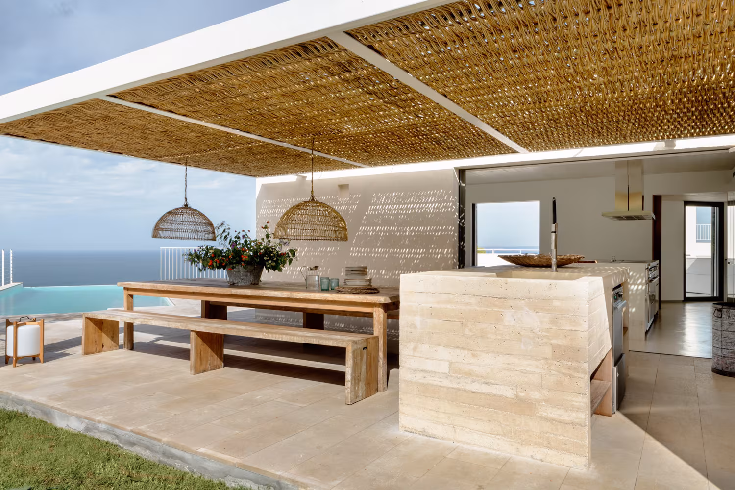 Cocina exterior con vista al mar, pérgola de cañizo e isla de obra revestida de travertino