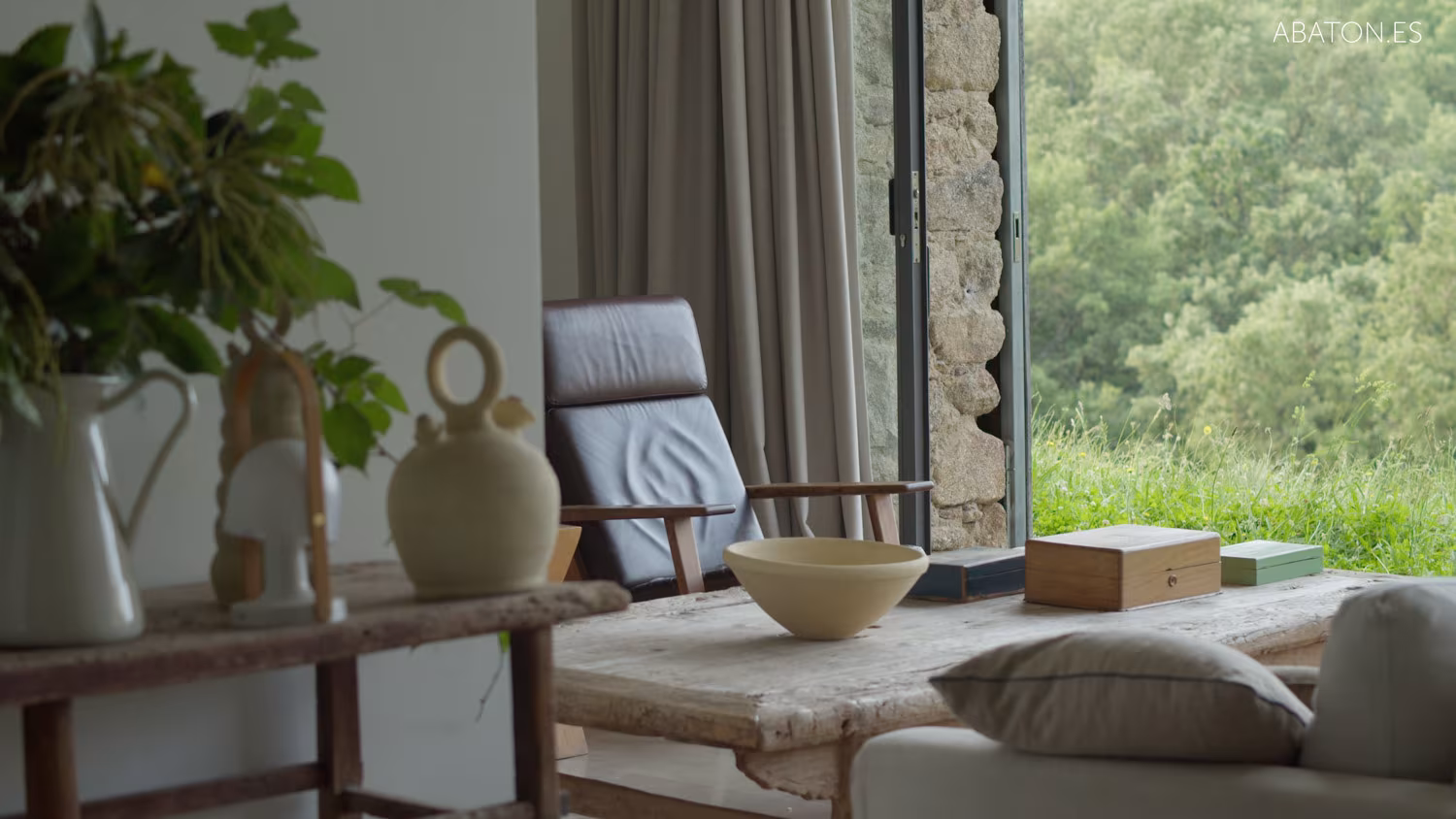 Rincón de estar con silla de cuero, mesa rústica de madera y ventanal de suelo a techo al jardín