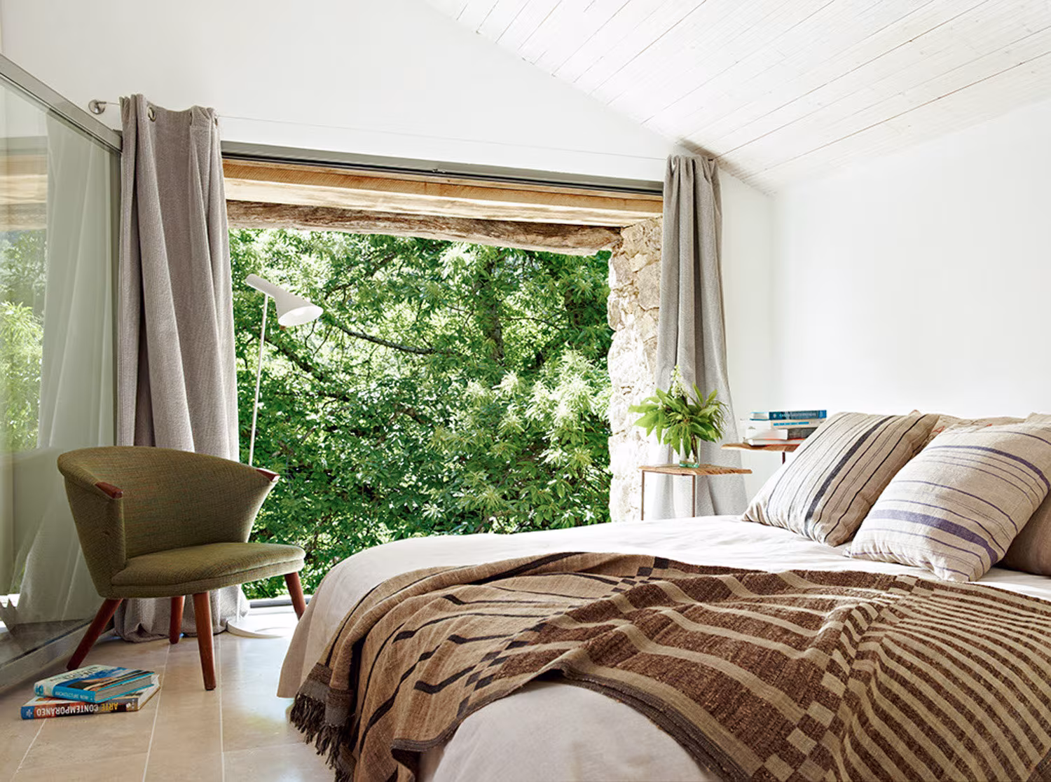 Dormitorio rústico con cama doble, manta de lino, cojines a rayas y ventanal al bosque