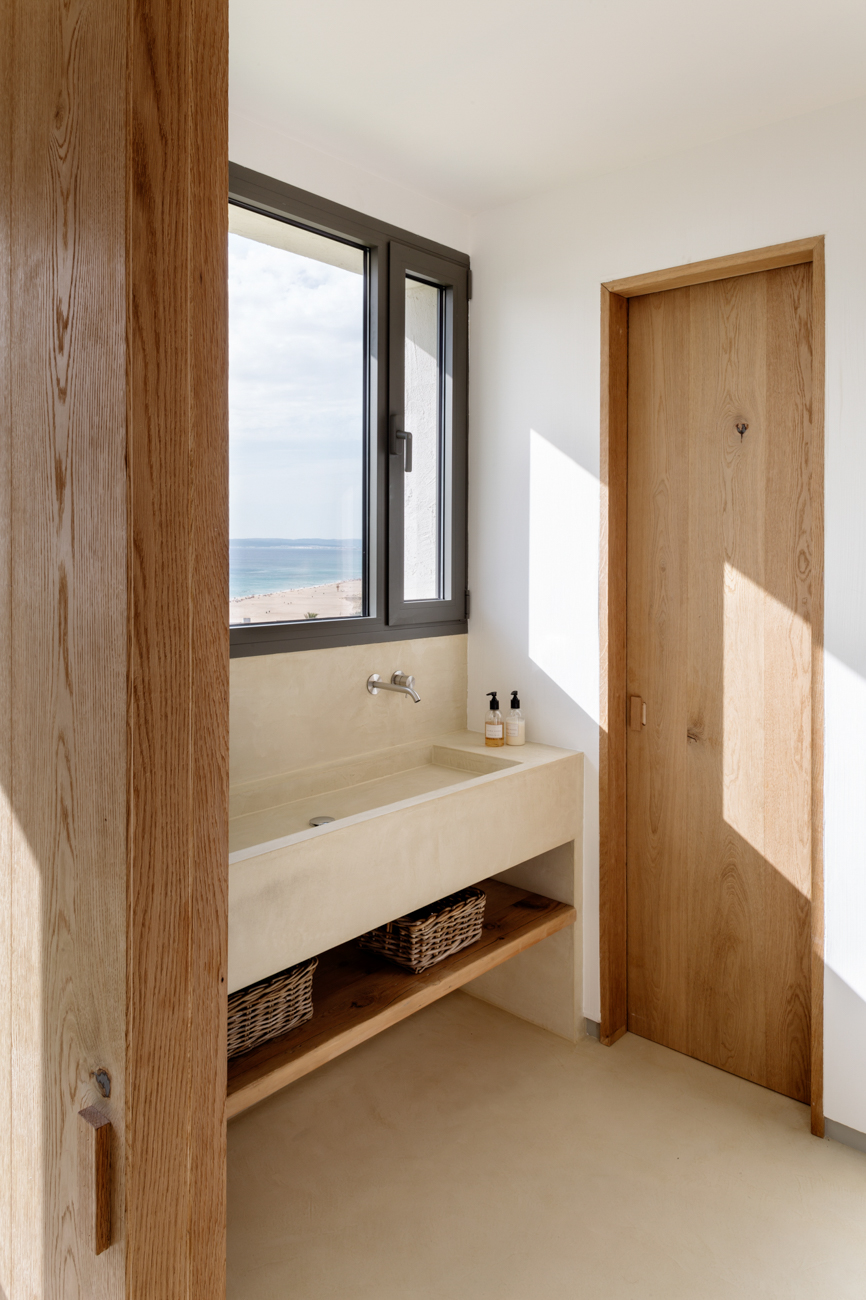 ABATON-proyecto-at-2 Baño minimalista con lavabo de microcemento en tonos beige, cesta de mimbre en estantería inferior y ventana con vistas al mar Mediterráneo, enmarcada por carpintería de madera natural.