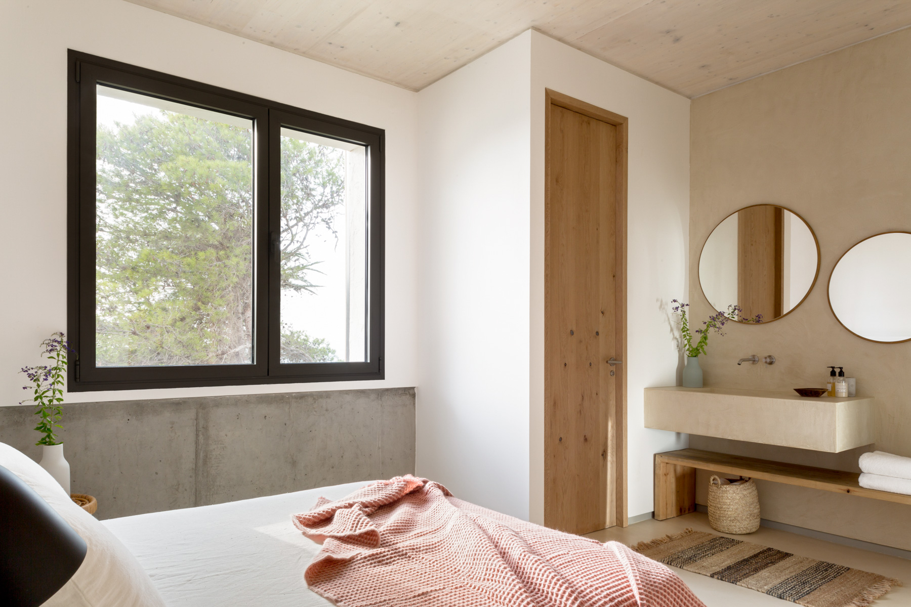 ABATON-proyecto-at-3 Dormitorio con baño integrado de estilo minimalista y natural. La habitación presenta una ventana negra con vista a árboles, paredes de yeso, madera clara y concreto visto. El baño incluye lavamanos de obra con espejo redondo y decoración con textiles suaves y fibras naturales, evocando calma y diseño mediterráneo.