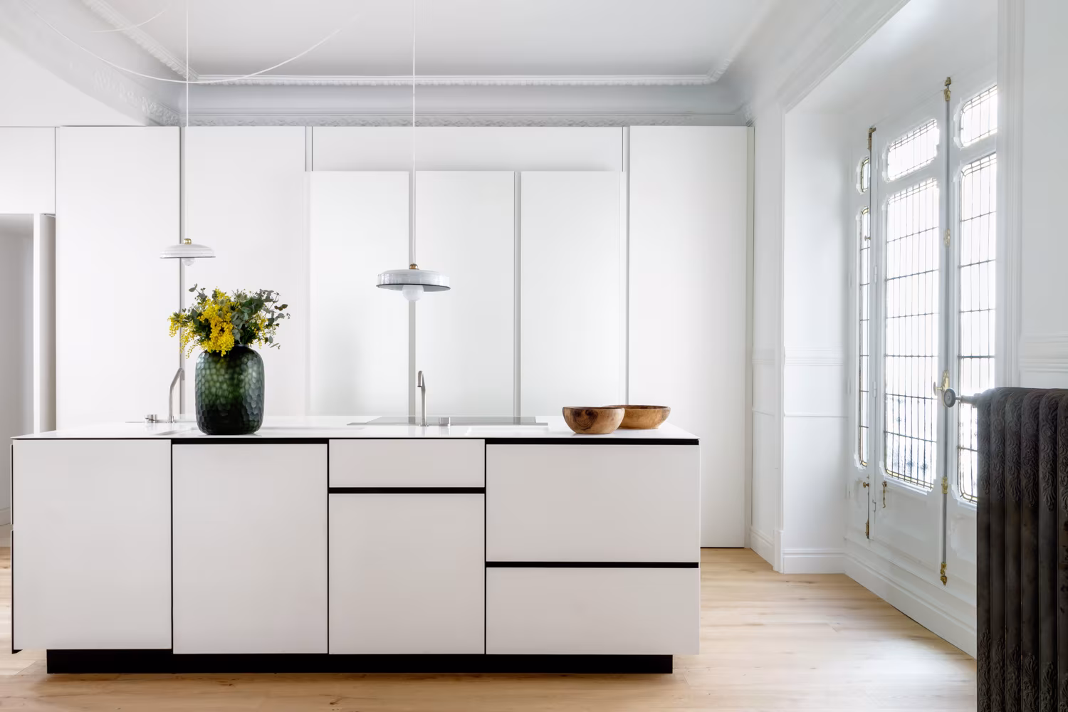 Cocina blanca tras reforma, con isla central minimalista, suelo de madera clara y molduras originales restauradas en el techo.
