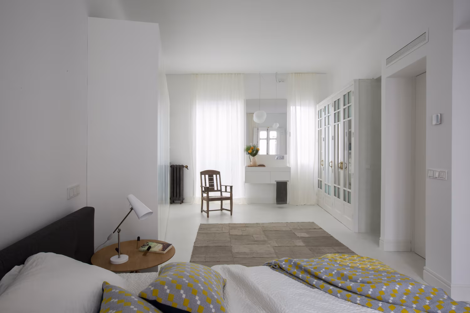 Dormitorio luminoso con cama doble, textiles geométricos, escritorio flotante con espejo y silla de madera sobre alfombra neutra