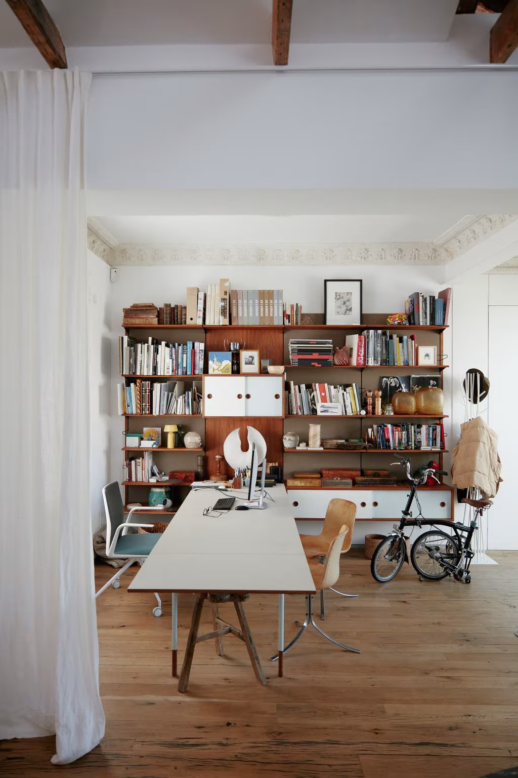 Study with a timber bookcase, long white table and wood-plank floor under a beamed ceiling