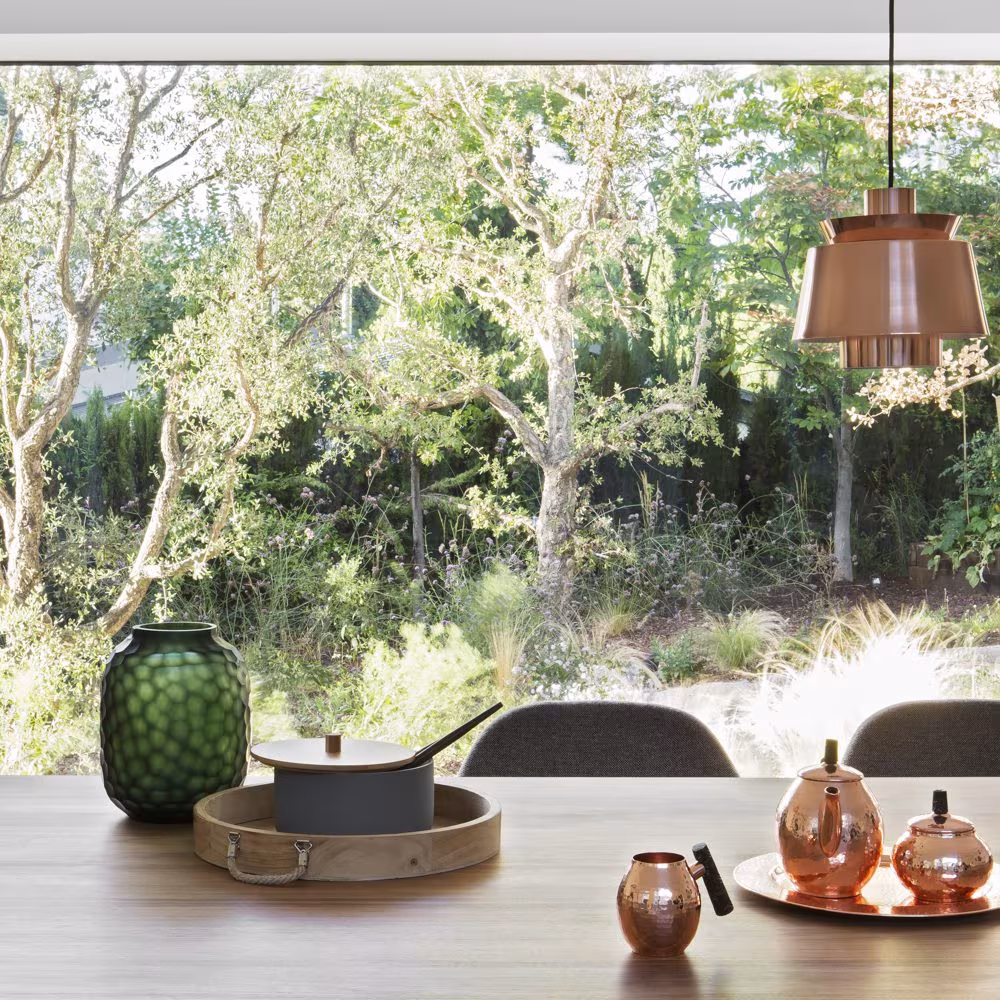 Mesa del comedor de Casa L con cobres y un jarrón verde frente al ventanal sobre el jardín arbolado