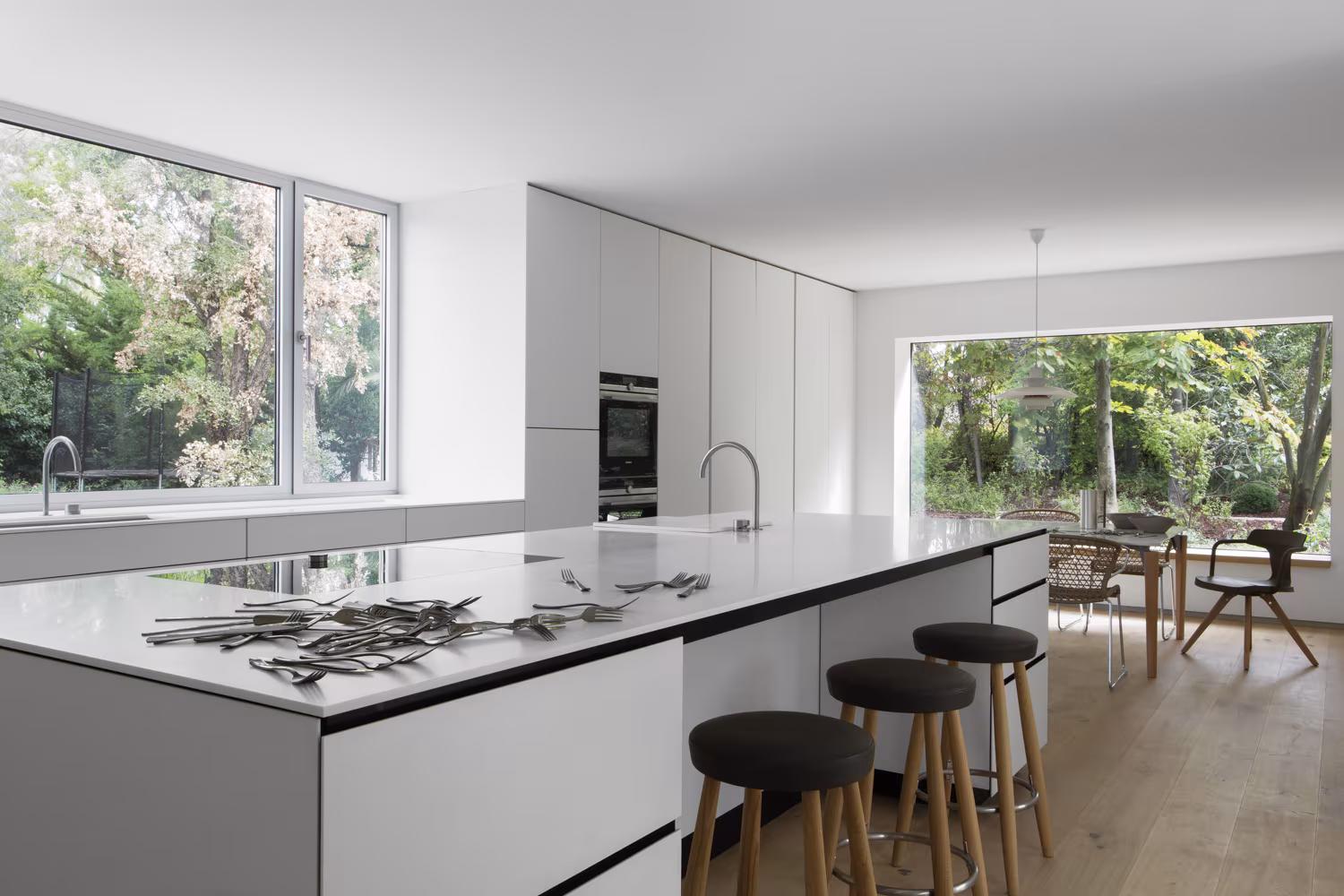 Cocina de Casa L con isla blanca de encimera negra, taburetes y ventanas al arbolado del jardín