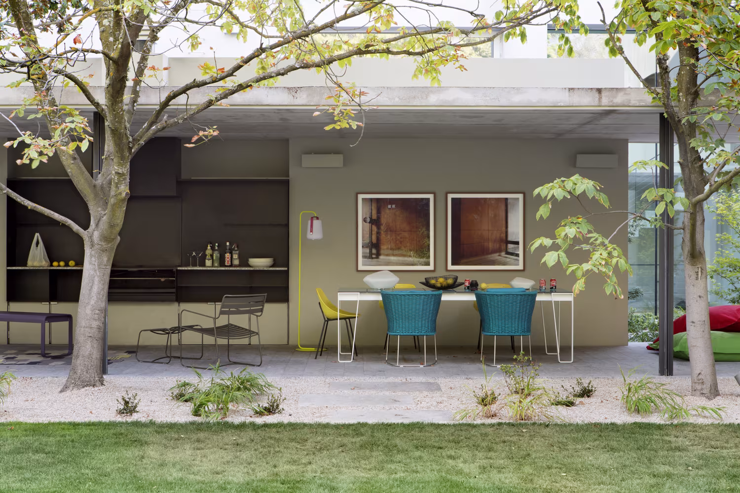 Porche abierto de Casa L con comedor de sillas de colores y cocina exterior entre los árboles del jardín