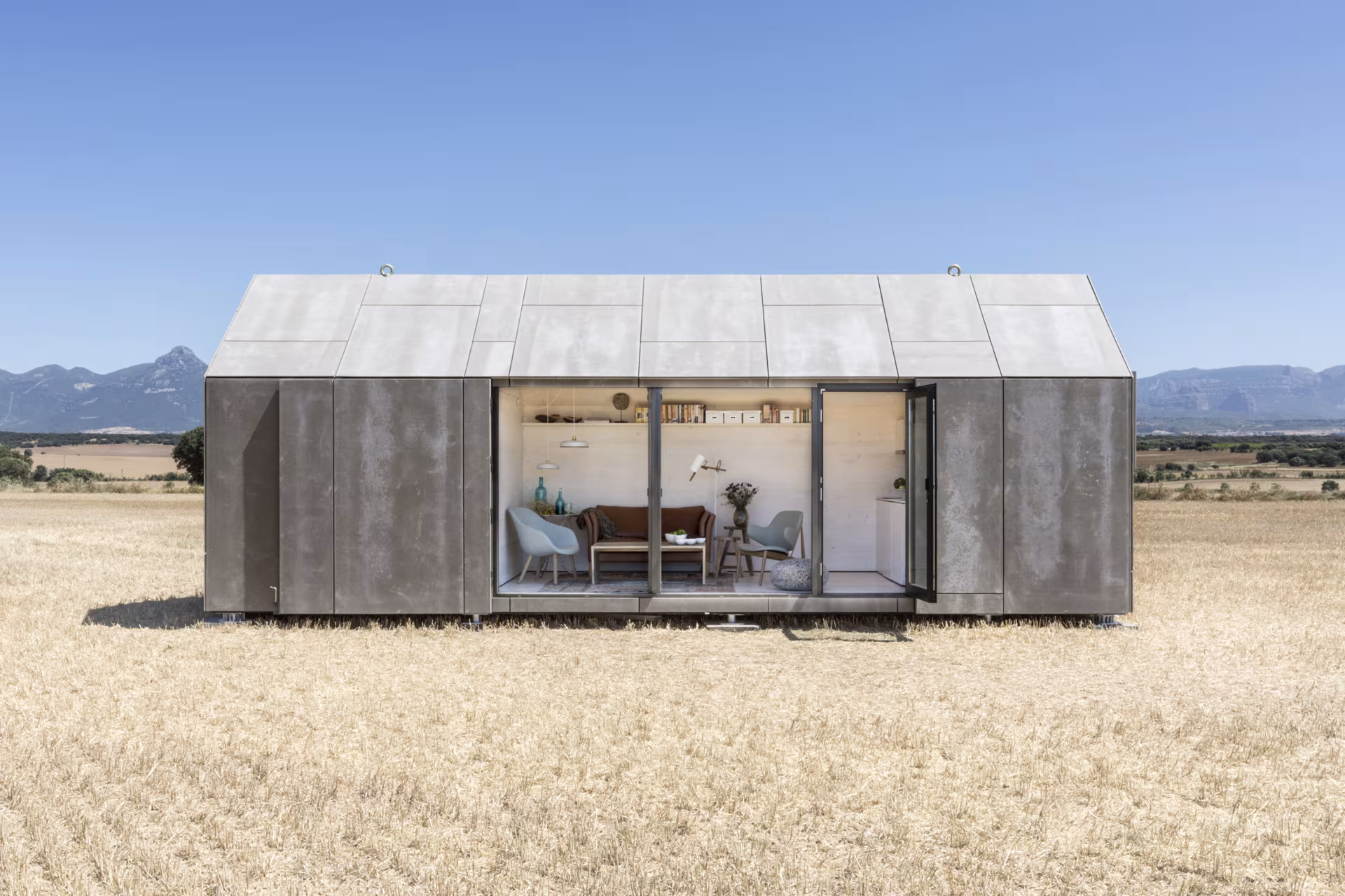 Casa transportable ÁPH80 vista de frente, con su salón iluminado tras la carpintería de vidrio y montañas al fondo