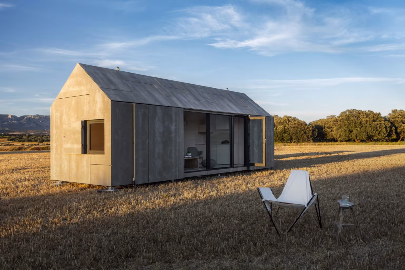 Casa transportable ÁPH80 revestida de madera y chapa oscura sobre el campo con una silla de tijera en primer plano