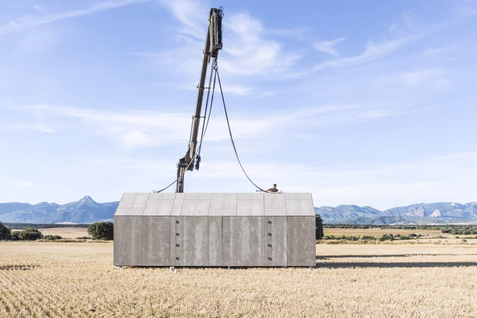 Casa transportable ÁPH80 izada por una grúa sobre el campo, con la sierra al fondo