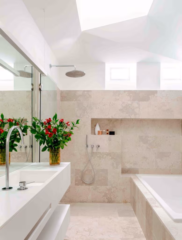 Baño de piedra clara con ducha, bañera y lucernario en la reforma integral del ático del Retiro, Madrid