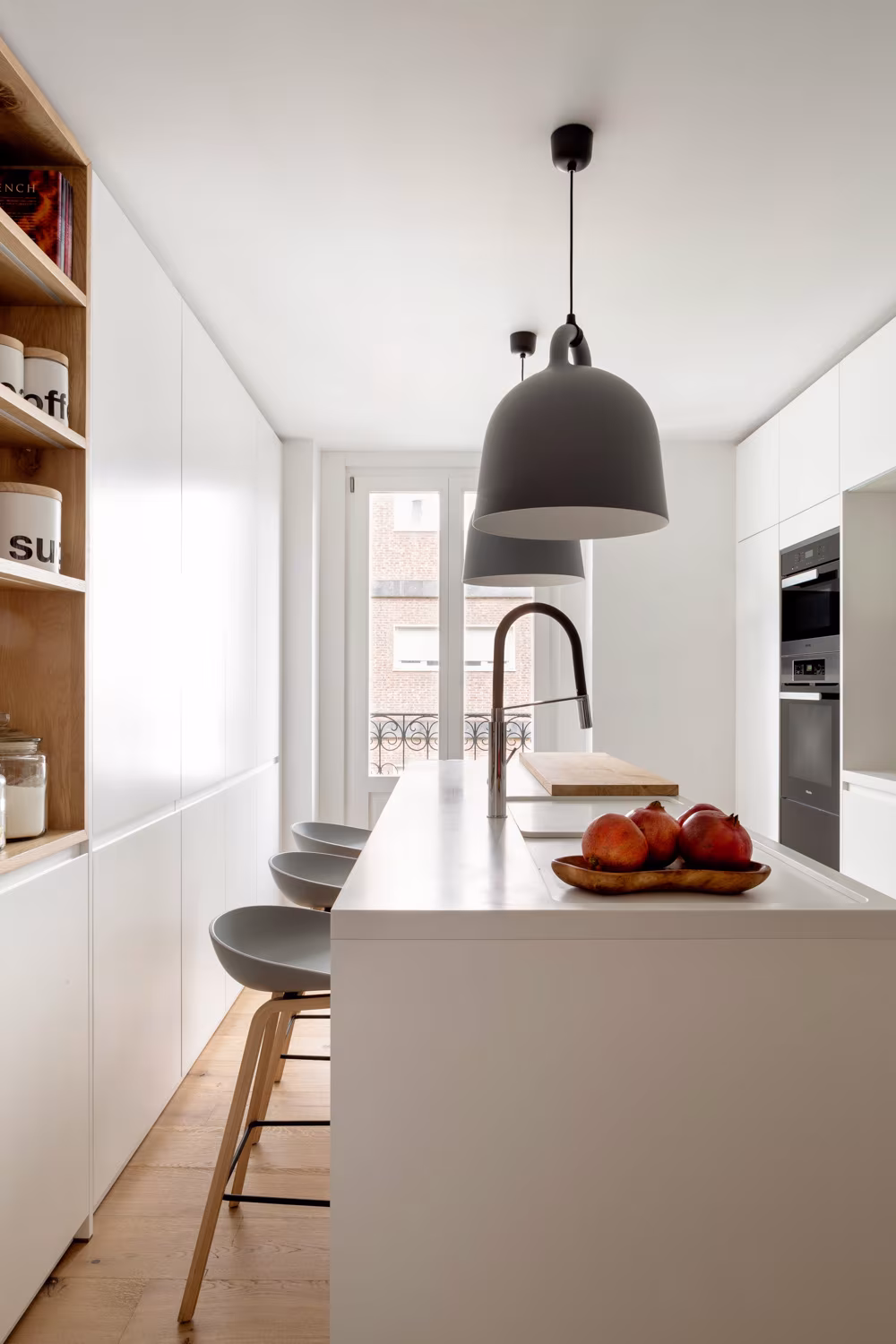 Cocina estrecha de Casa Ayala II con isla, taburetes y lámpara gris sobre muebles blancos y estantes de madera