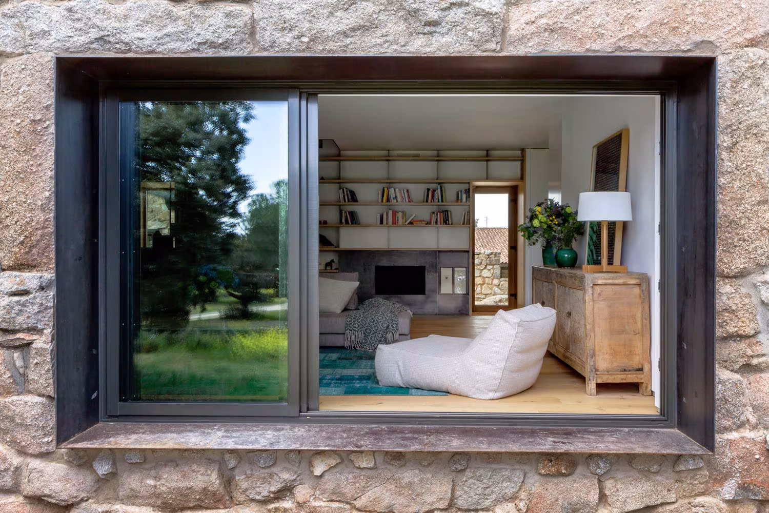 Ventana de marco metálico oscuro en un muro de granito que enmarca el salón de Casa de Campo LS de Ábaton