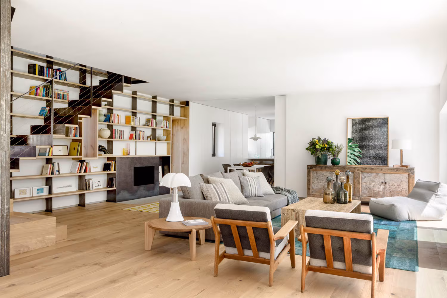 Salón de doble altura de Casa de Campo LS con biblioteca de madera y metal, butacas de roble y sofás grises