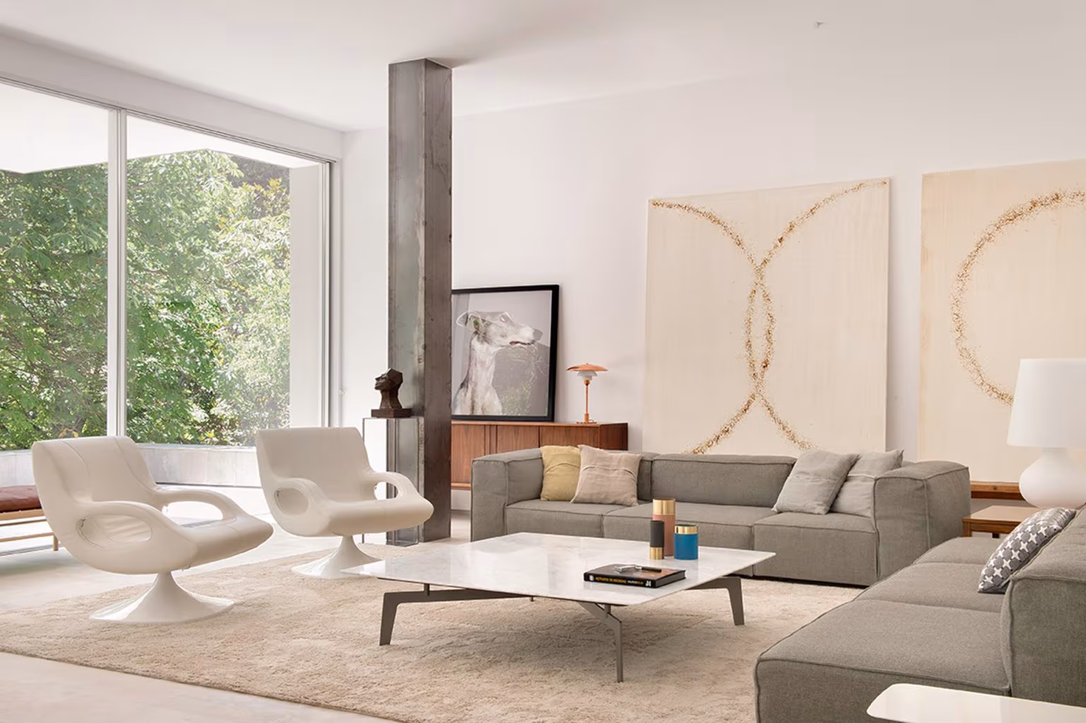 Salón de Casa L con sofá gris, pilar de acero, butacas blancas y dos grandes cuadros claros ante el jardín