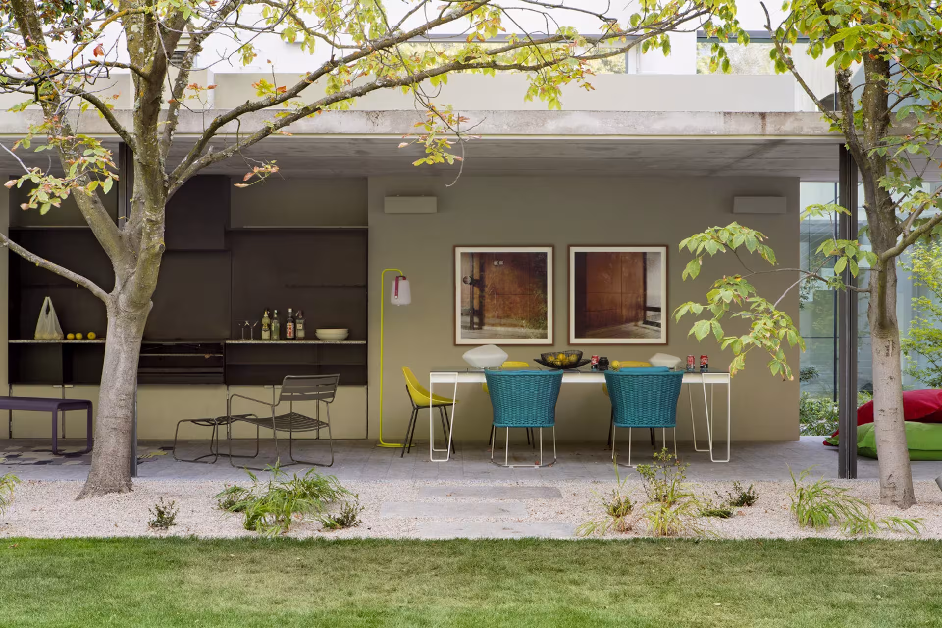 Porche con cocina exterior, mesa con sillas de colores y césped bajo un árbol en la Casa L, Madrid