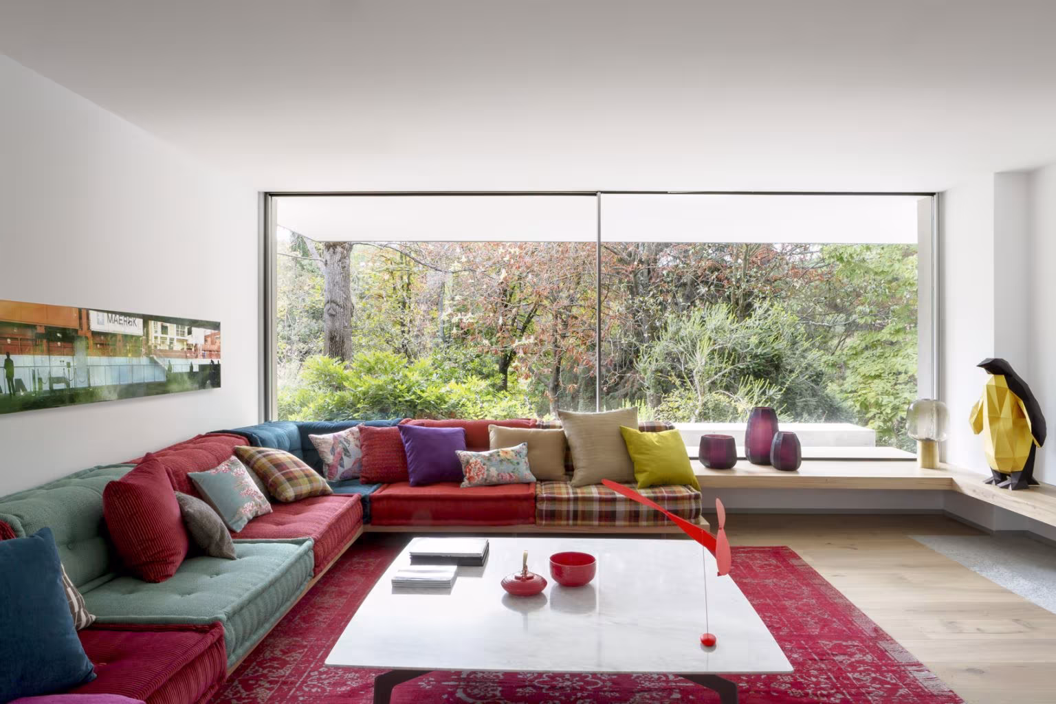 Salón de Casa L con sofá rinconero de colores, alfombra roja y un gran ventanal sobre el jardín