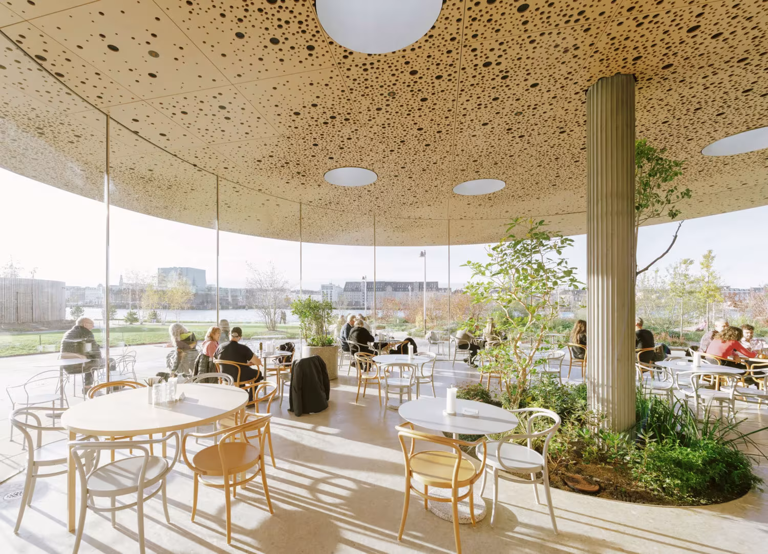 Cafetería bajo una gran cubierta de madera con mesas redondas y un pilar rodeado de plantas, Opera Park