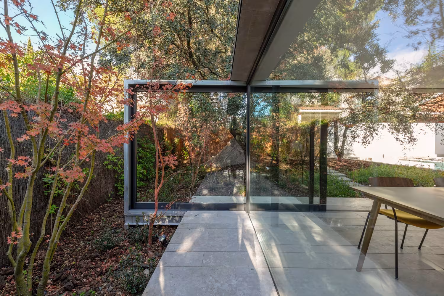 ABATON-pabellon-del-jardin-03 Glazed corner of the pavilion with timber table, red-leaved trees and pale stone paving