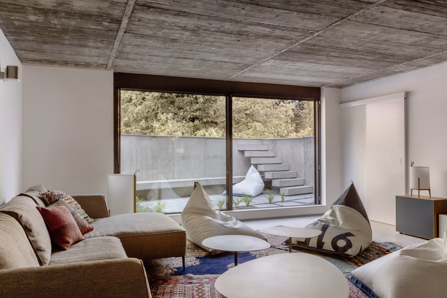 Sala de estar inferior con techo de hormigón, sofá y puffs junto a un ventanal sobre un patio, Casa de las luces