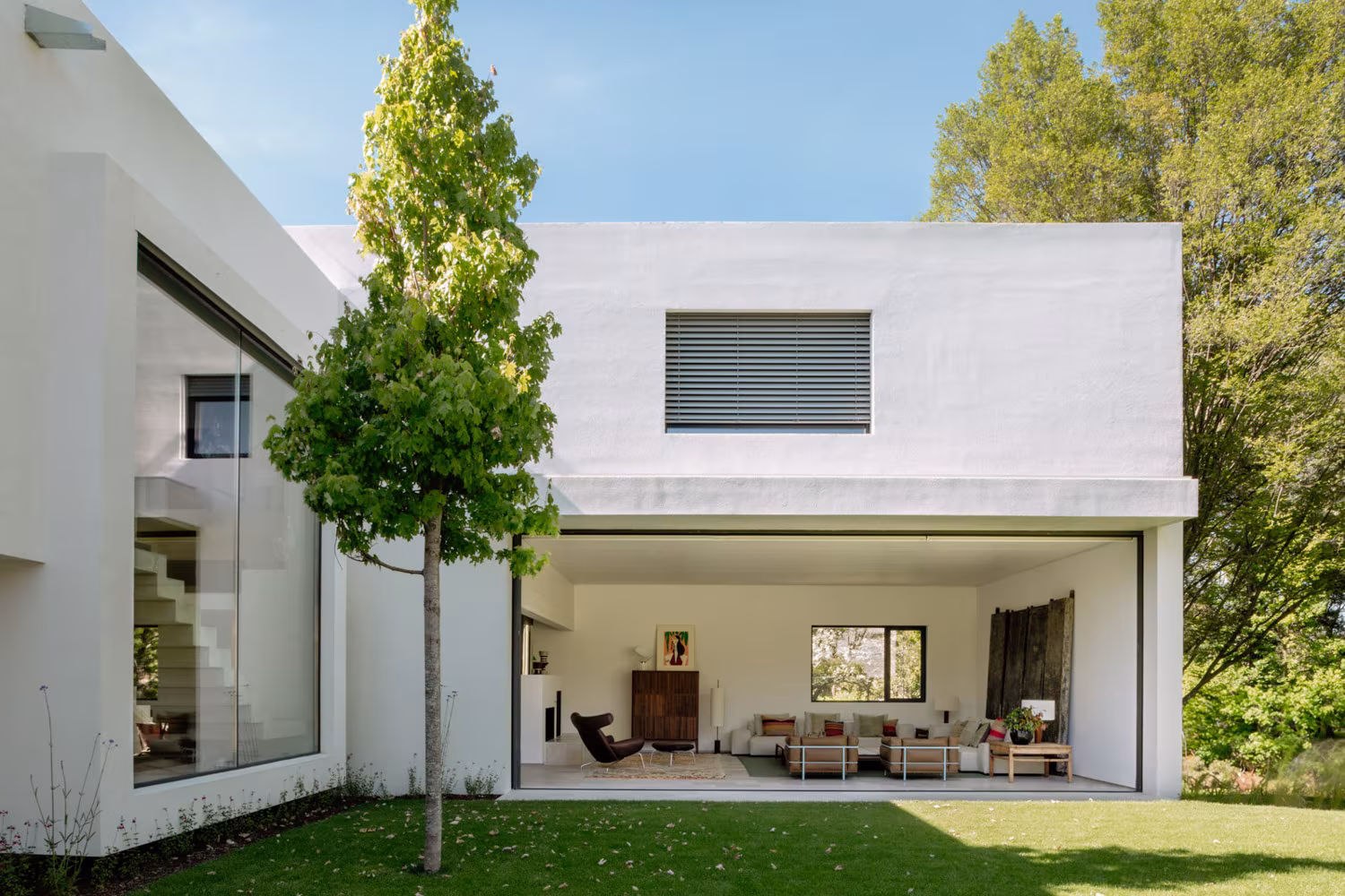 Volumen blanco con un gran hueco acristalado hacia el jardín y un árbol joven, Casa de las luces
