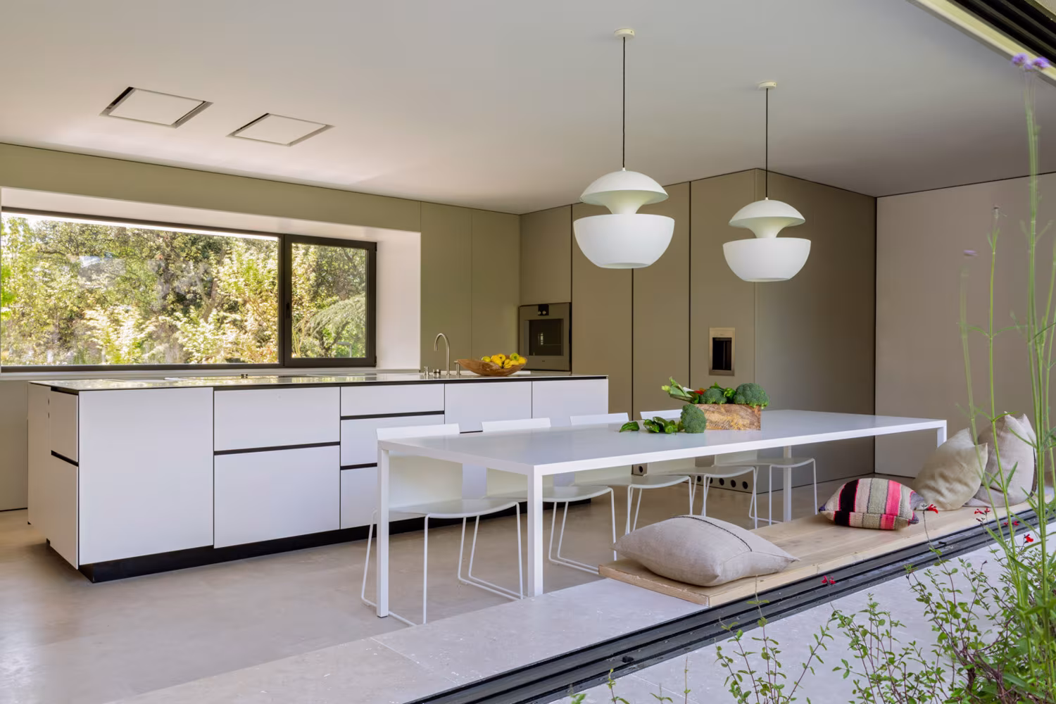 Cocina con isla blanca y mesa corrida bajo lámparas esféricas, con ventana hacia el bosque, en la casa de las luces