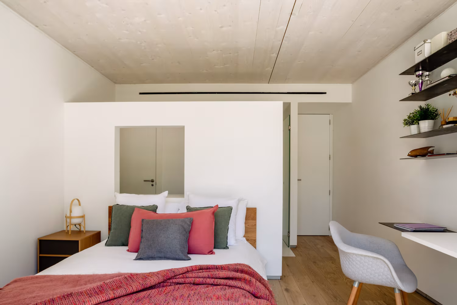 Dormitorio de techo de madera clara con cabecero bajo, cojines coral y escritorio, en la casa de las luces