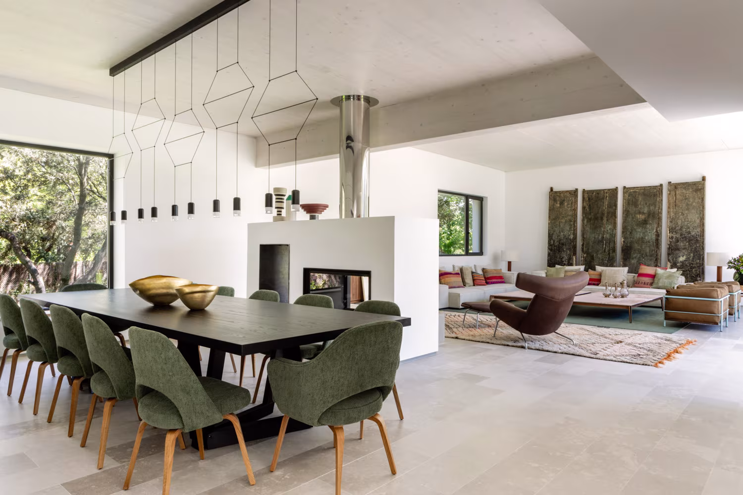 Comedor con mesa oscura, sillas verdes y lámpara geométrica junto a una chimenea de doble cara, Casa de las luces
