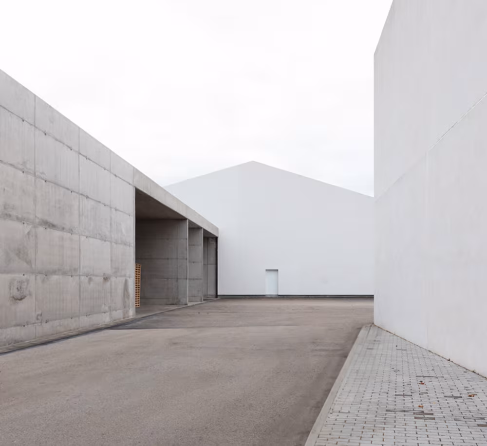 Muros de hormigón visto y volúmenes blancos a dos aguas alineados a lo largo de un patio pavimentado