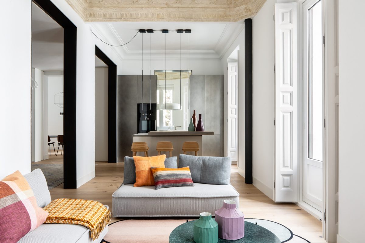 ABATON-reforma-almagro-1 Salón luminoso de apartamento con molduras clásicas y grandes ventanales con decoración colorida: sofás bajos y cojines vibrantes. Vista a cocina abierta moderna con isla central y taburetes de madera