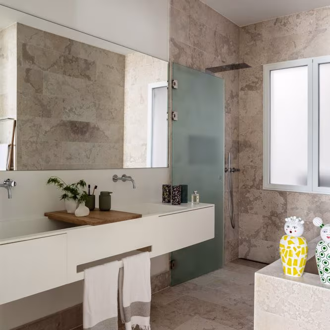 Baño de piedra con doble lavabo suspendido, mampara de vidrio verde y ventana en la reforma de Almagro de Ábaton