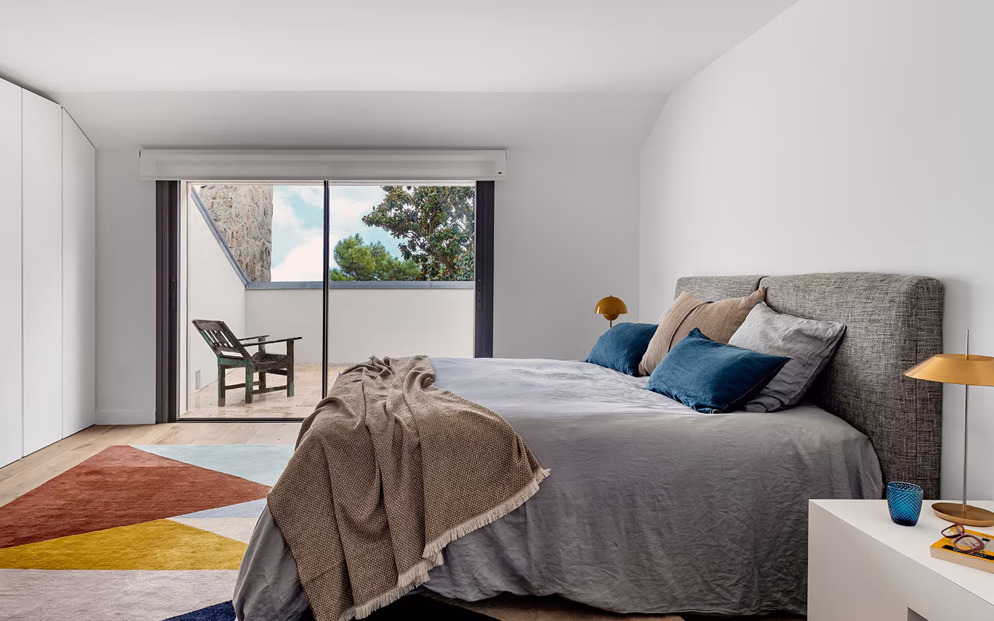 Dormitorio luminoso con cama gris, cojines azules, manta beige, alfombra multicolor y acceso a terraza con silla exterior
