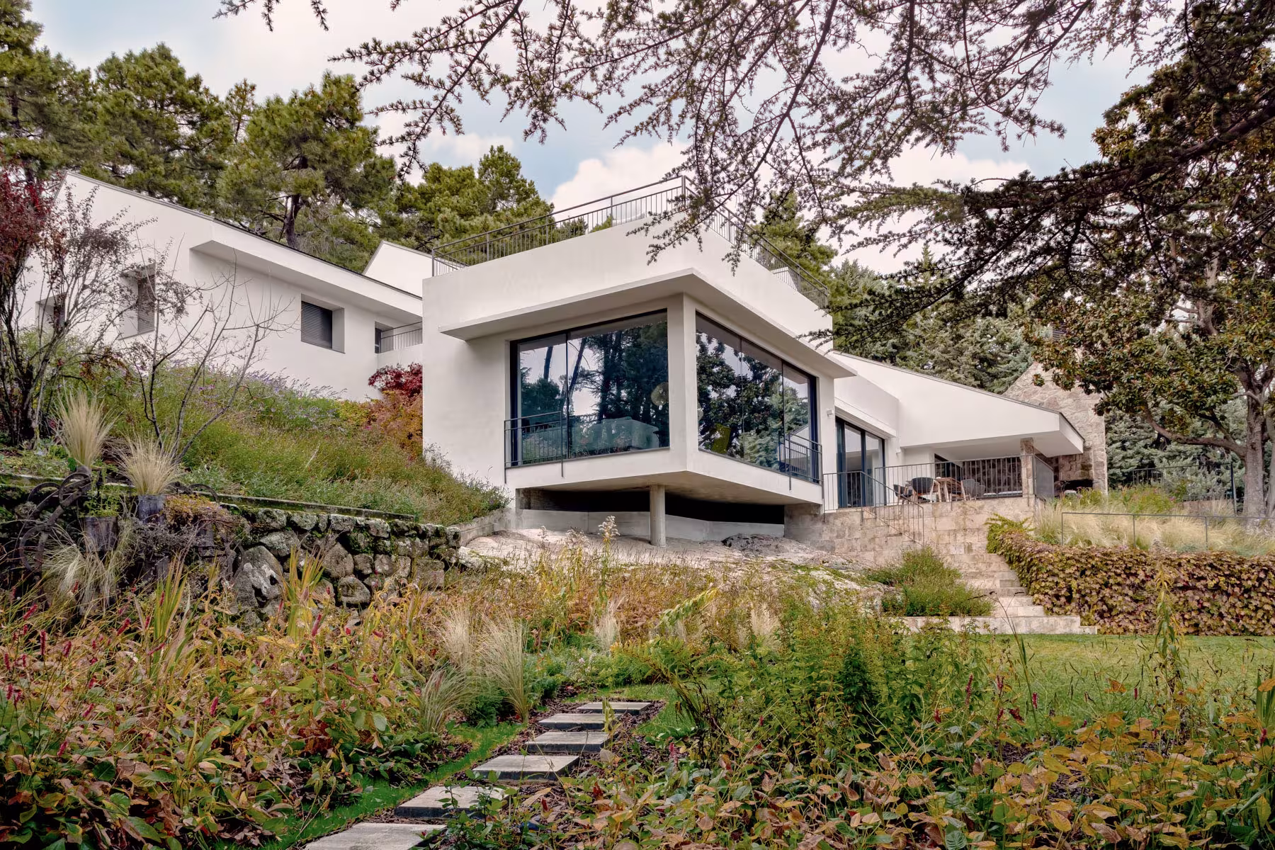 Fachada de casa moderna blanca en la montaña, rodeada de vegetación y grandes ventanales con vistas al jardín.