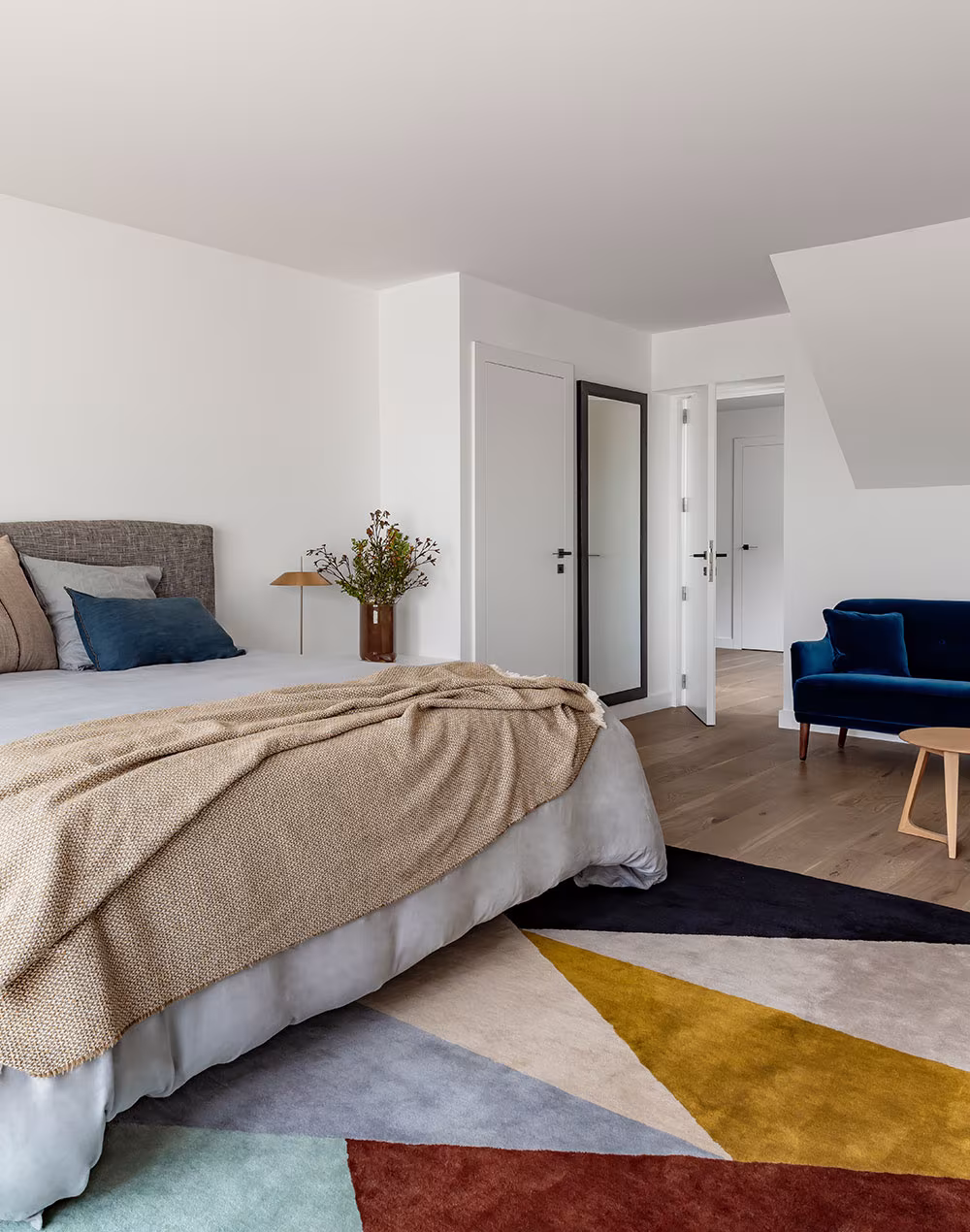 Dormitorio con cabecero tapizado, alfombra geométrica de colores y butaca azul de terciopelo