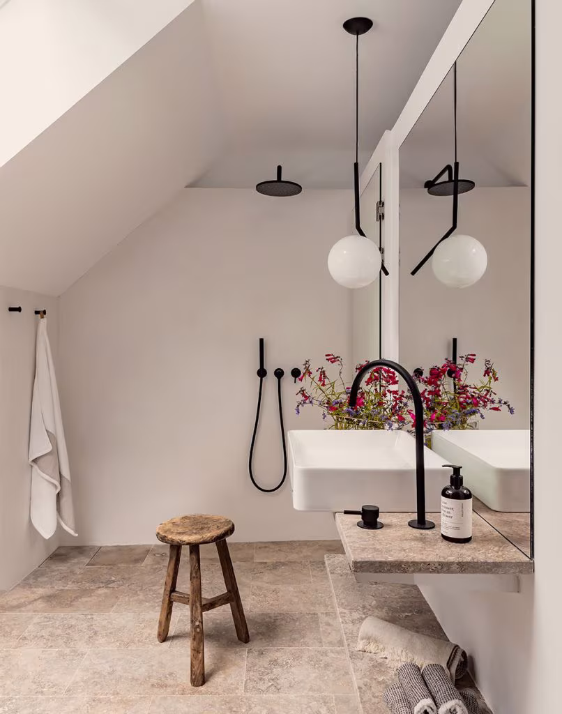 Baño minimalista con lavabo suspendido, grifería negra, banco rústico, flores frescas y suelo de piedra natural beige