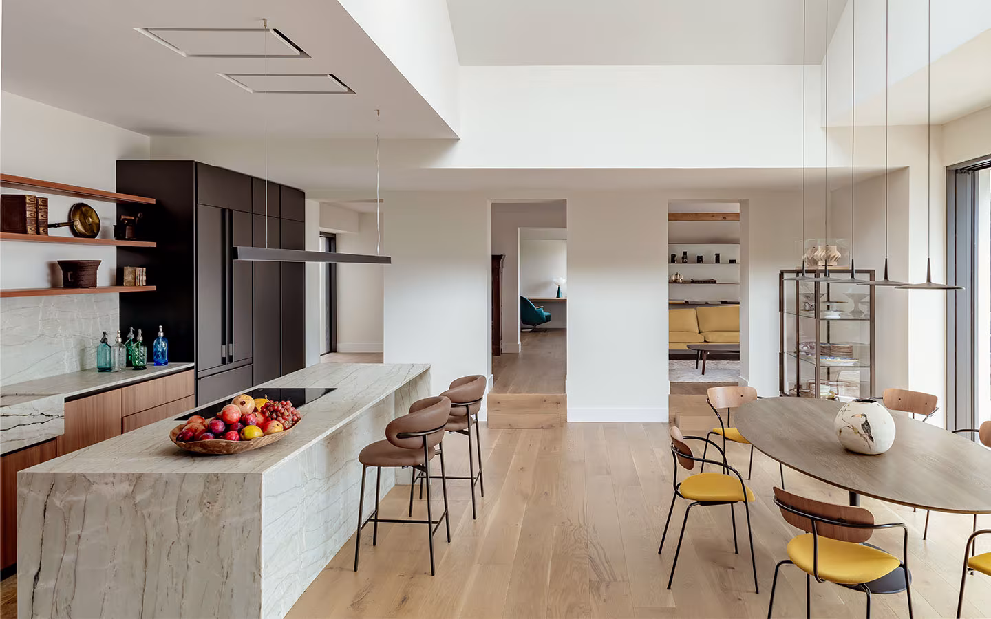 Cocina moderna con isla de mármol, taburetes altos, comedor con sillas amarillas y suelo de madera clara que aporta calidez.