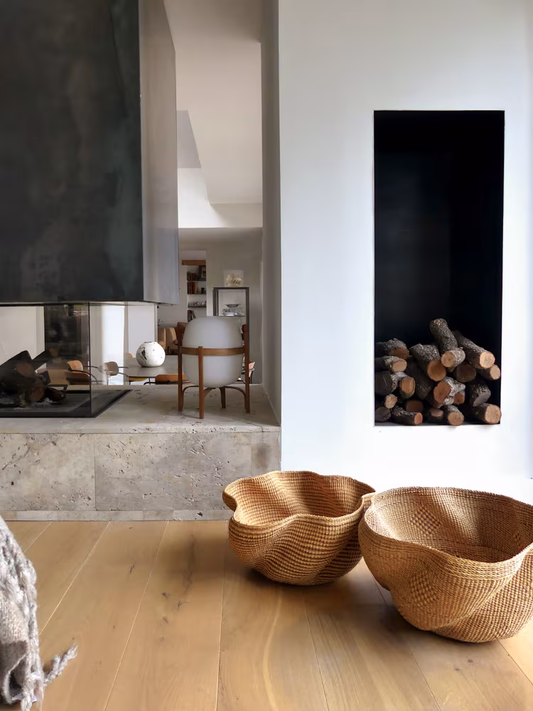 Interior con chimenea moderna de piedra, leña apilada y cestas decorativas sobre suelo de madera clara