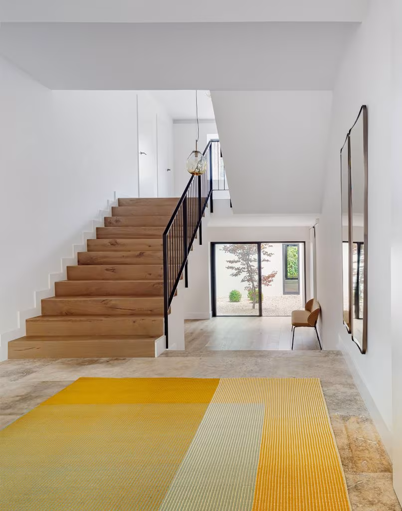 Recibidor moderno con escalera de madera, alfombra amarilla, silla tapizada y grandes ventanales al patio