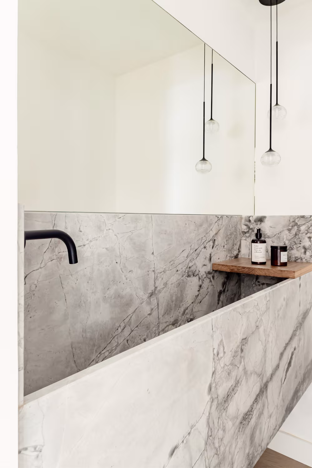 Bañera moderna de mármol gris con grifería negra mate y estantería de madera, iluminación colgante minimalista y espejo de gran formato